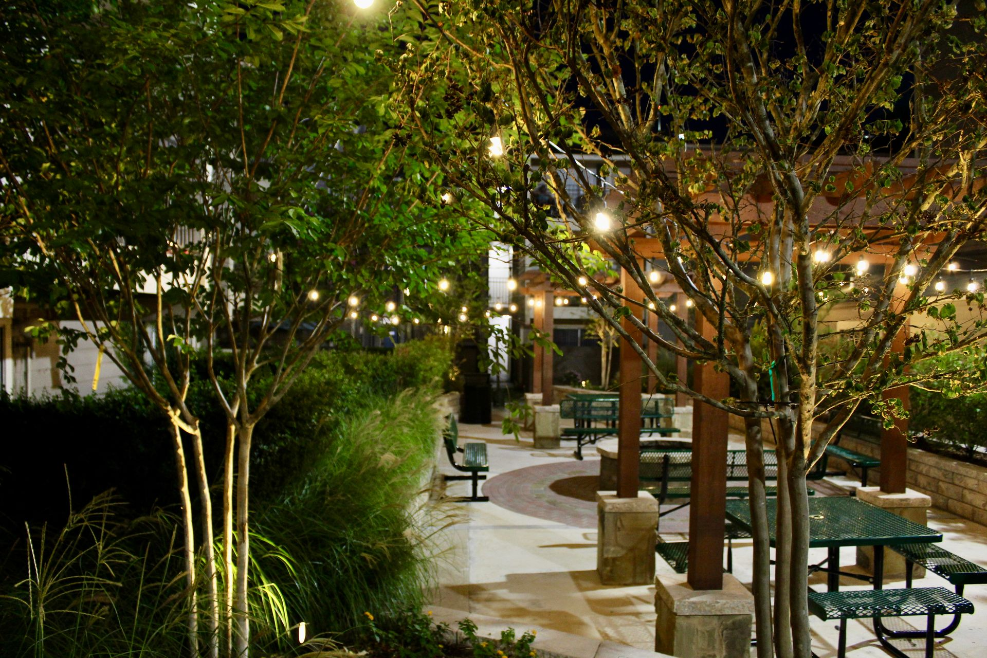 Outdoor dining area at night, string lights, picnic tables, trees, brick and stone pathways.