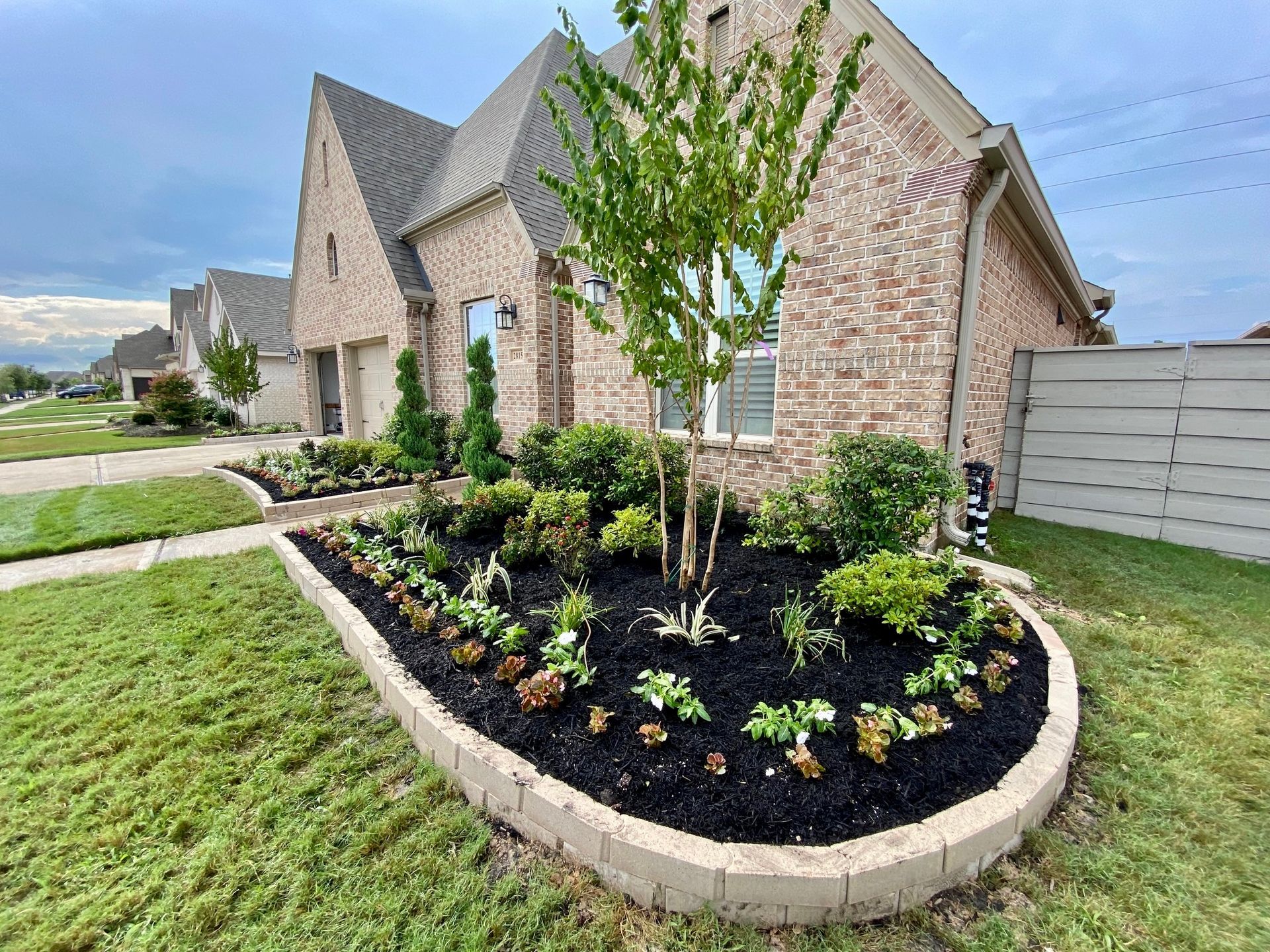 Brick house with a landscaped front yard bed, edged with tan blocks, under a cloudy sky.