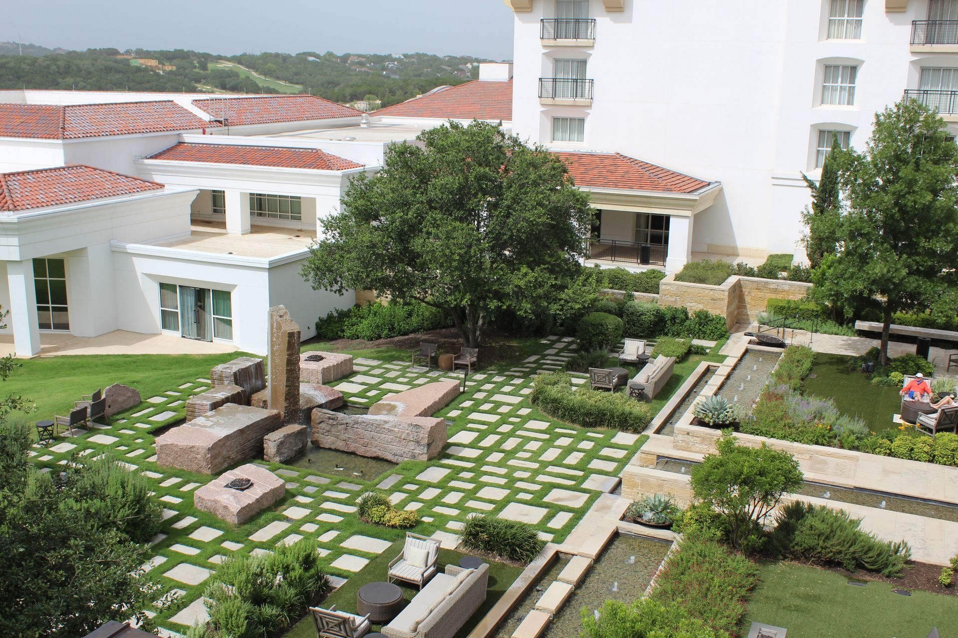 Hotel courtyard with stone sculptures, grass, and water features. White buildings with red tile roofs.
