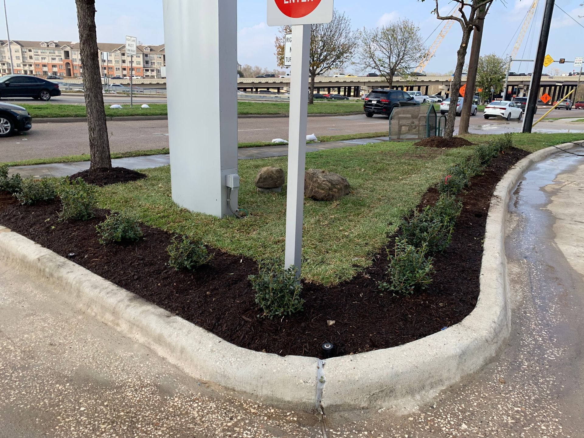 Corner curb with landscaped bed, green grass, shrubs, brown mulch, and a large white sign.