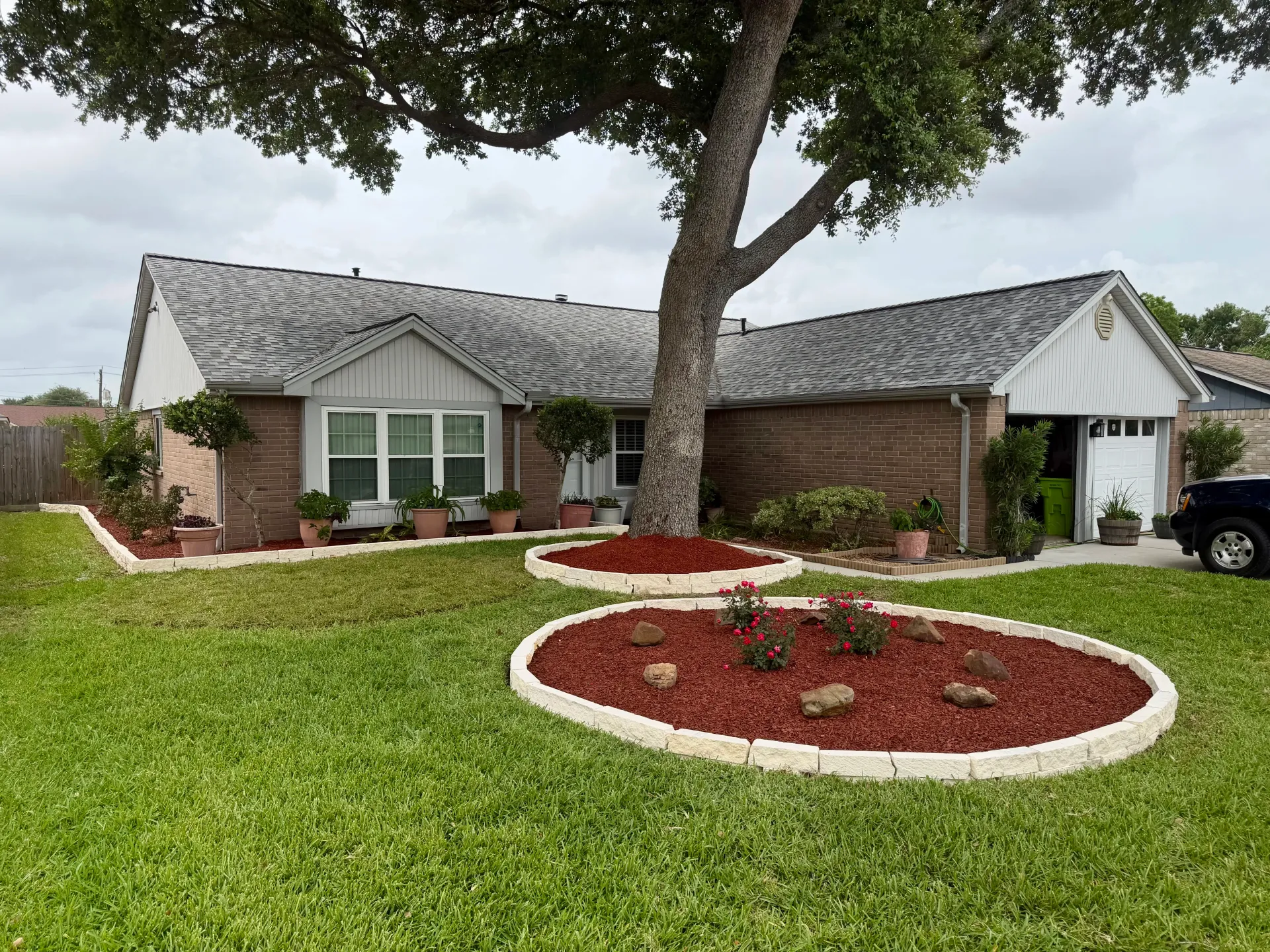 Single-story suburban house with manicured lawn, circular flower beds, and a large tree in front