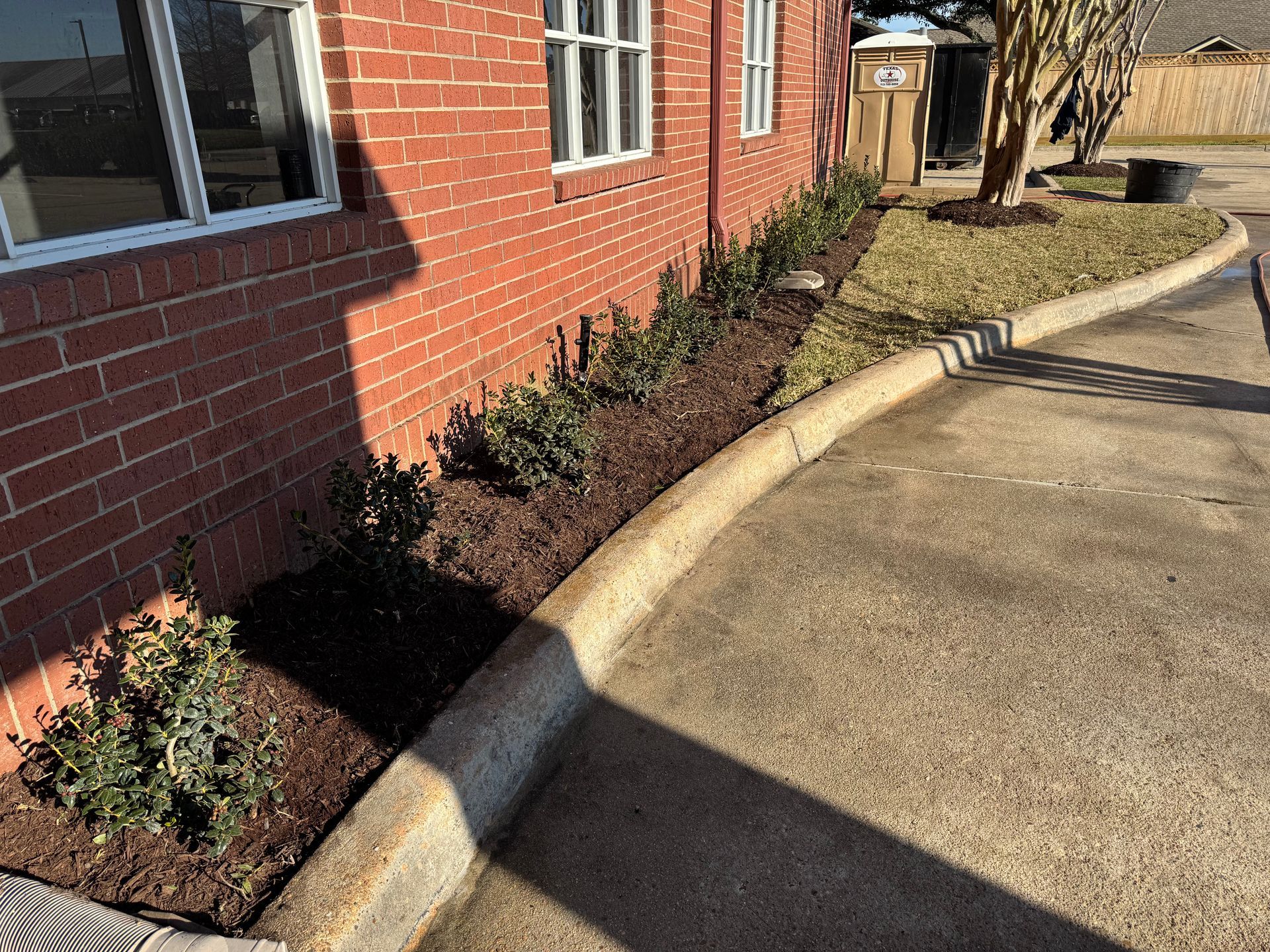 Exterior of a brick building with a gravel bed, black fencing, and a wet, paved area in the foreground.