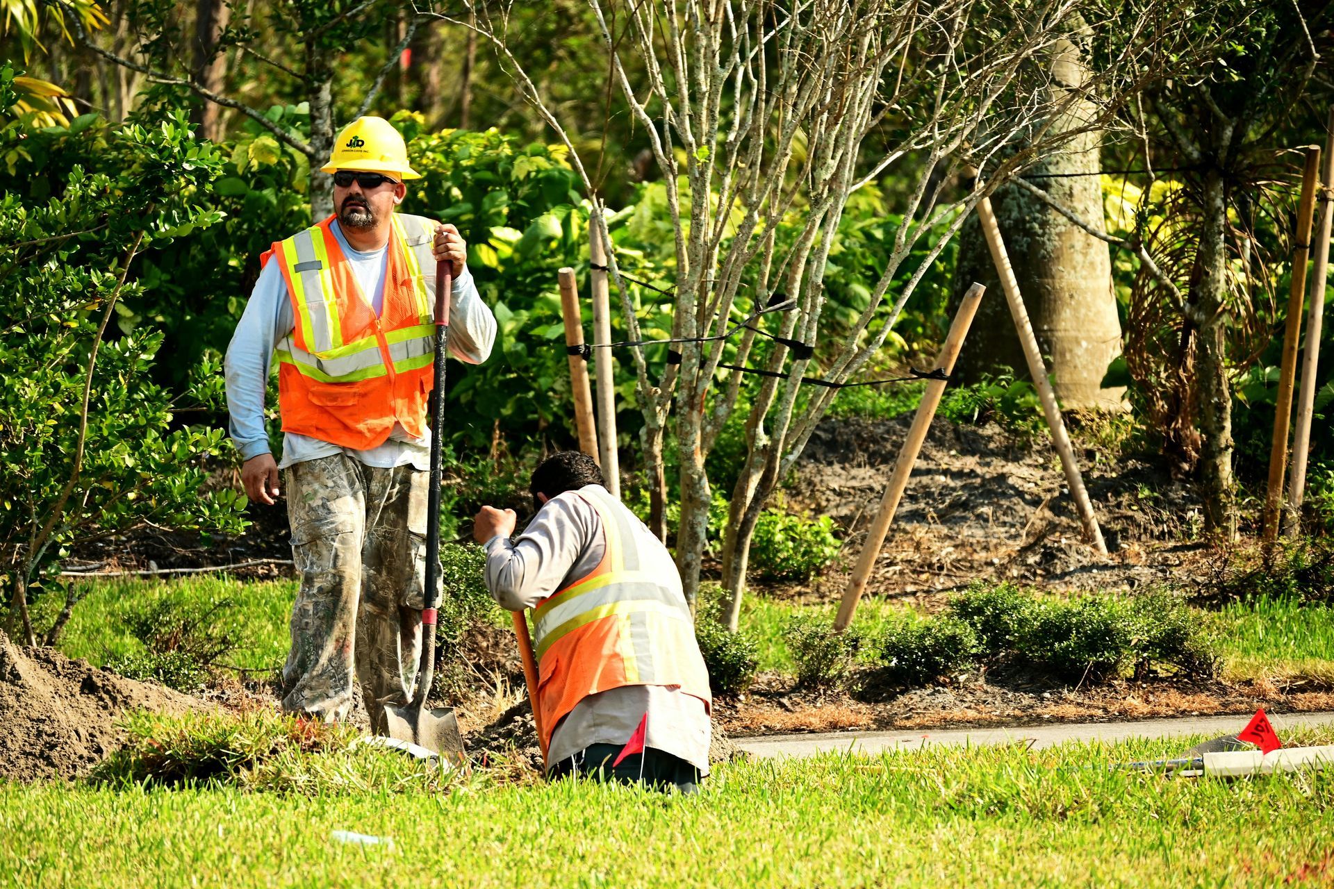 Two construction workers in safety vests and hard hats work outdoors near young trees.