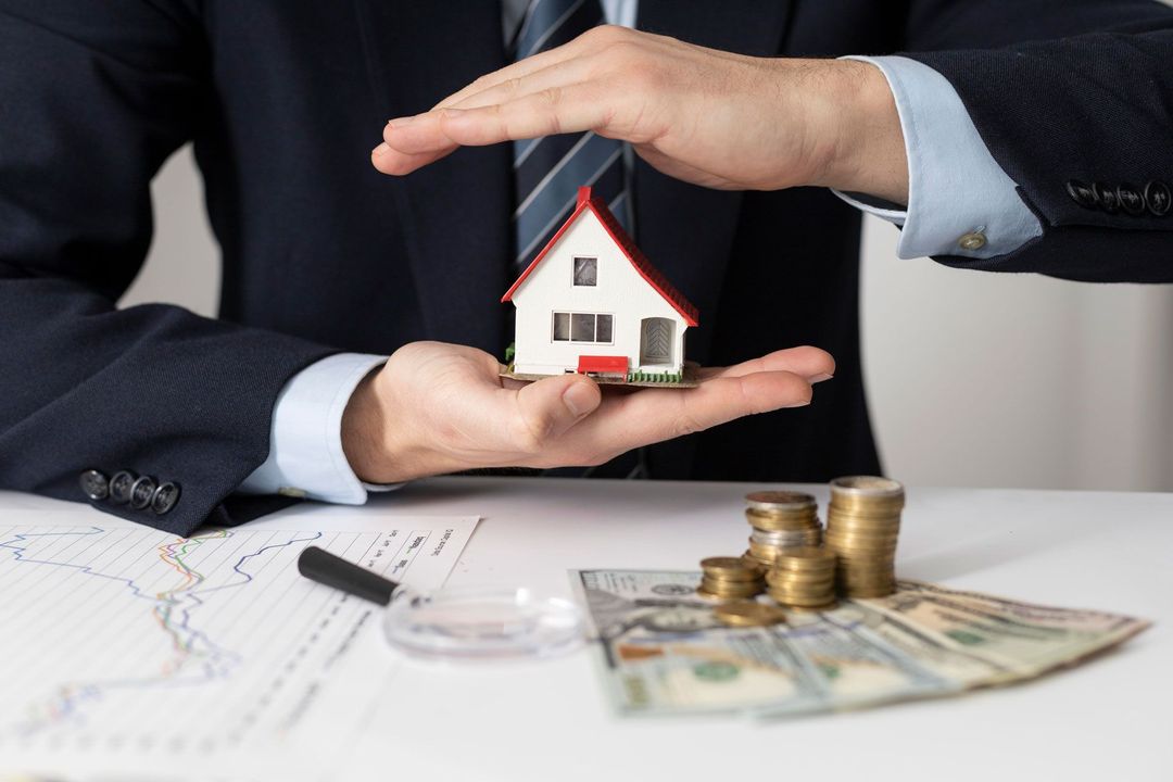 A man in a suit holding a model house with his hands, protecting it. Money and coins are visible on the table.