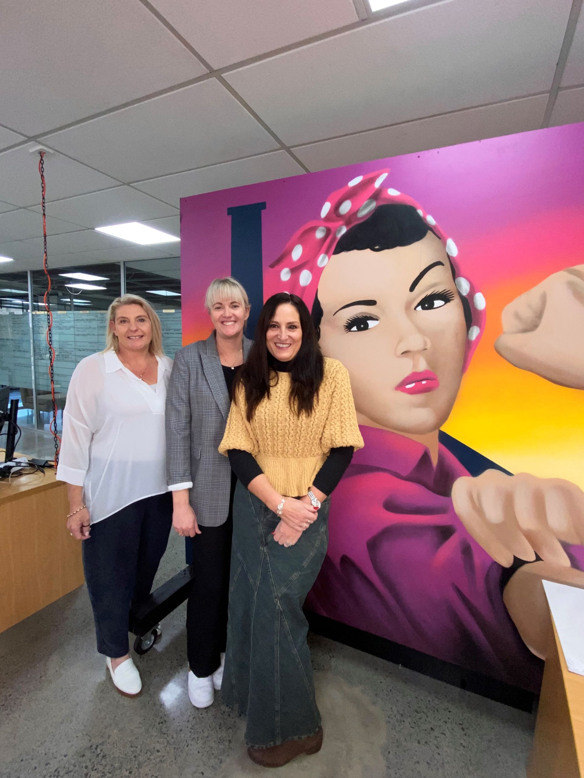Three women are standing in front of a mural of a woman.