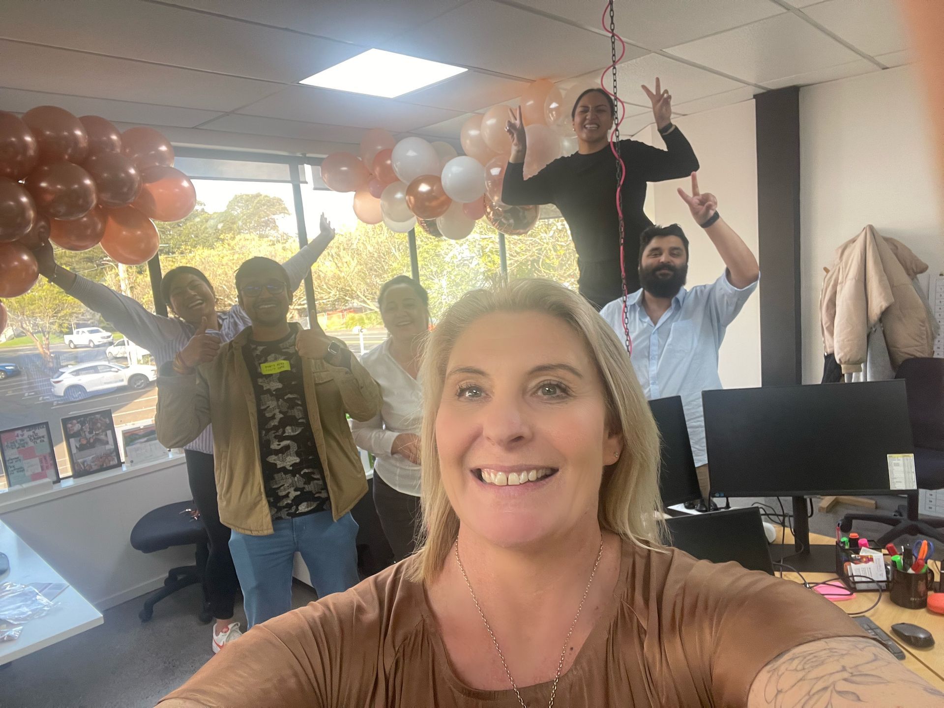 A group of people are taking a selfie in an office with balloons hanging from the ceiling.