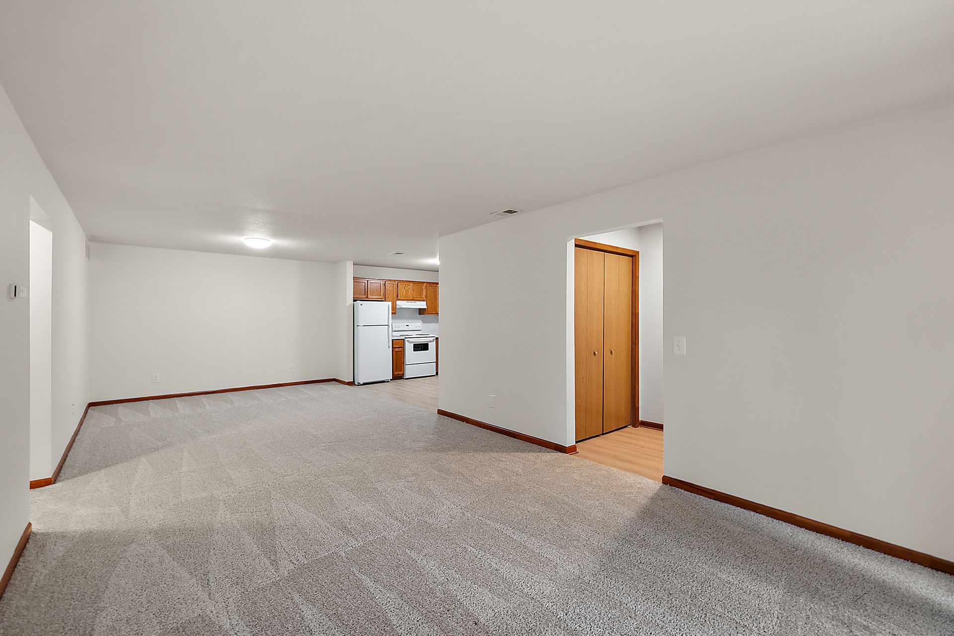 An empty room with a carpeted floor and a kitchen in the background.