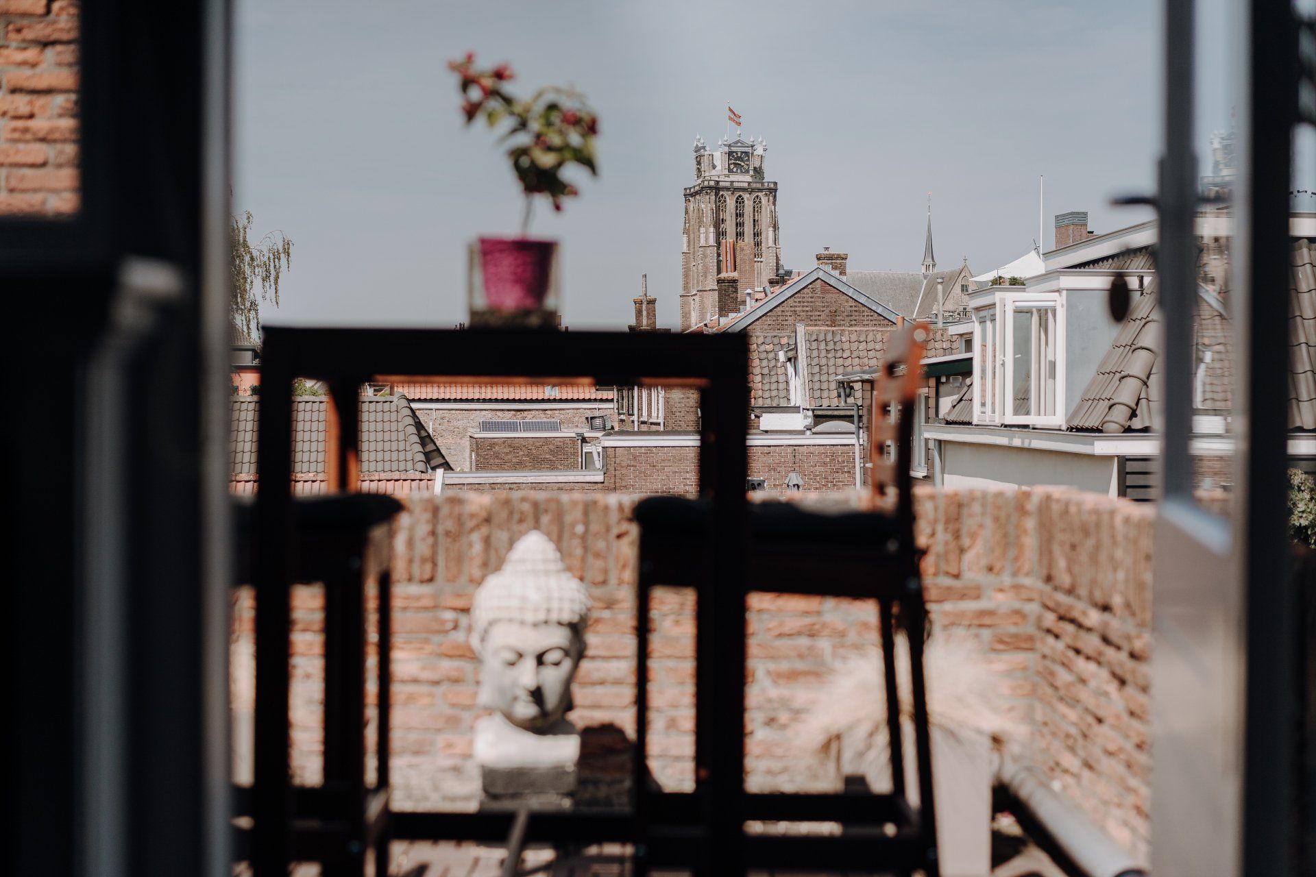 Uitzicht vanaf het balkon: Boeddhabeeld, tafel met plant, daken van de stad en kerktoren. Zonnige dag.