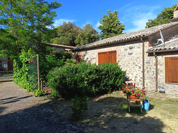 Cortile di un cottage in pietra con persiane in legno, cespugli verdi e fiori in vaso sotto un cielo azzurro brillante.