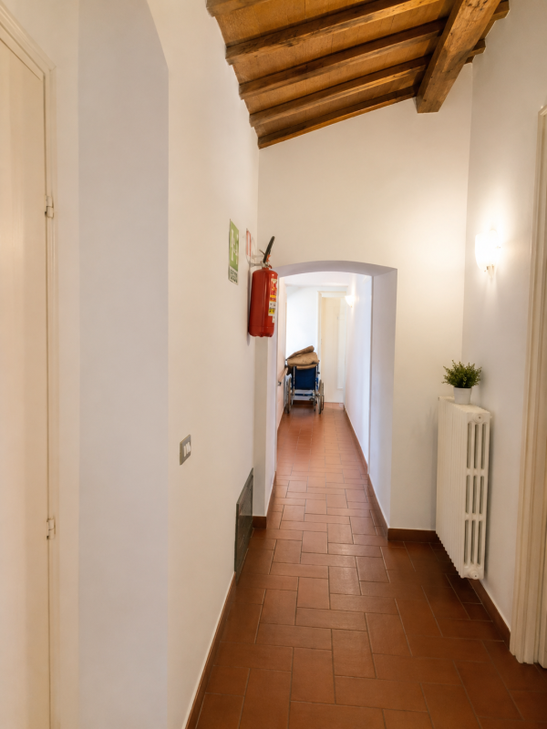Lungo e stretto corridoio con pavimento in piastrelle di terracotta, pareti bianche, soffitto con travi in legno e una sedia in fondo.