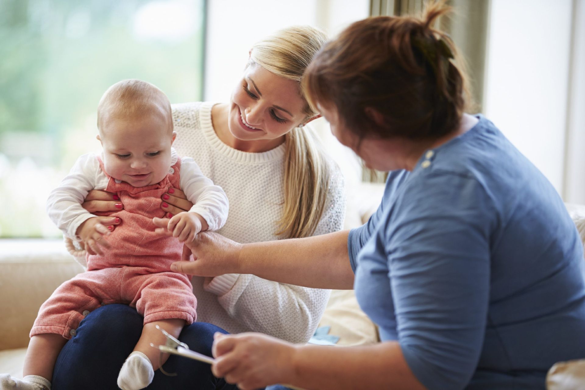 A professional assesses a seated infant on a parent's lap, checking the baby's foot with a small instrument in a room.