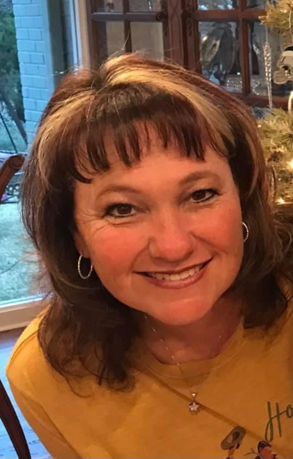 A woman in a yellow shirt is smiling in front of a christmas tree.