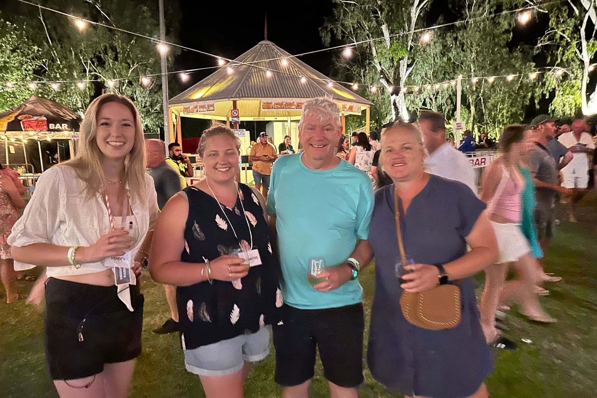 Four people smiling at a night festival, gazebo in background, string lights, others mingling.
