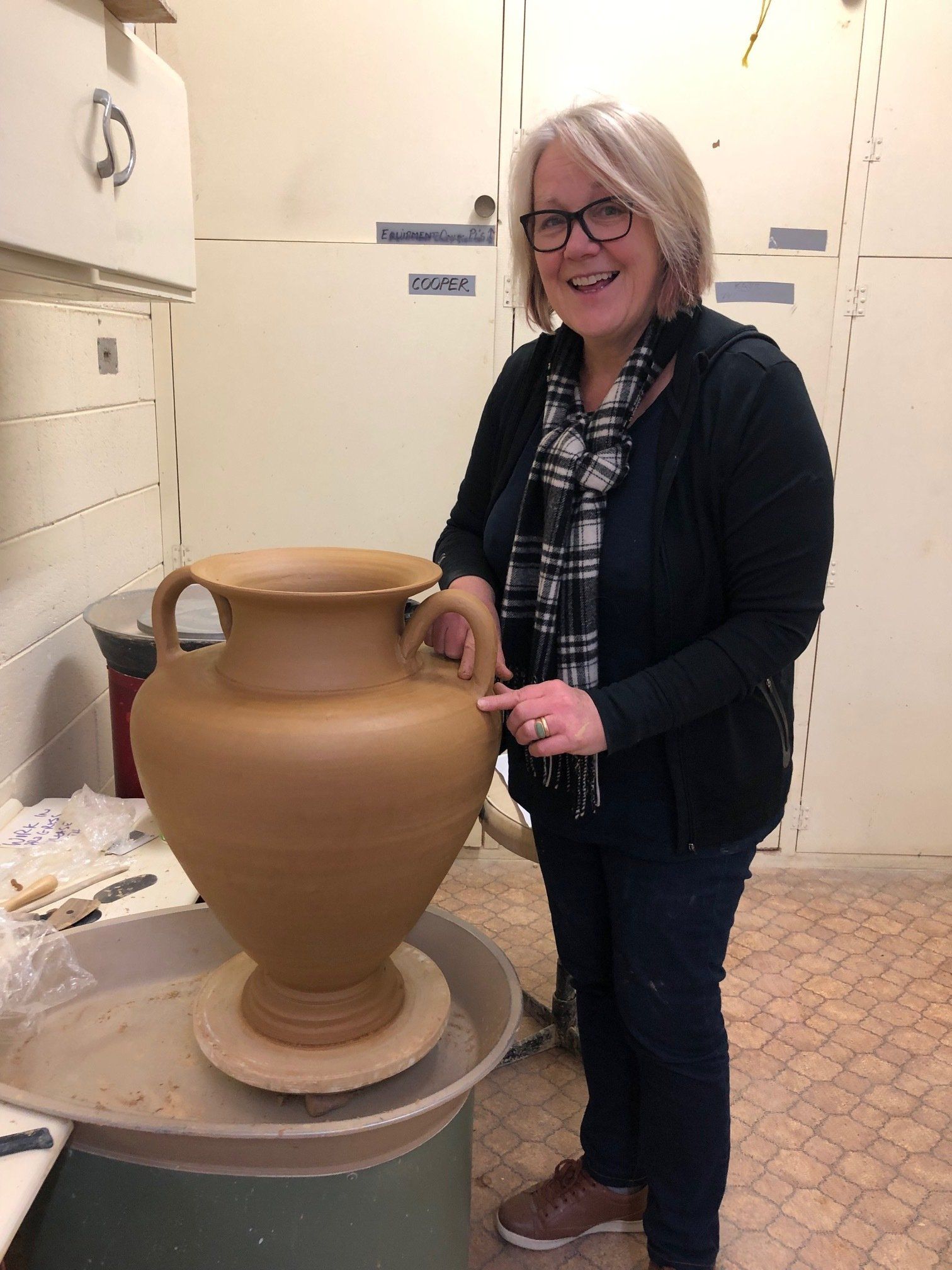 A woman is standing next to a large vase on a pottery wheel.