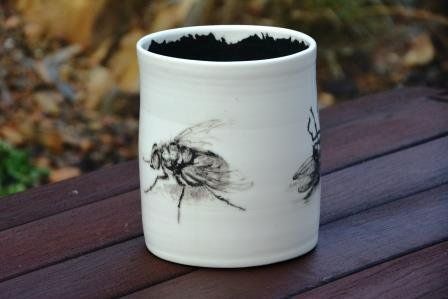 A white cup with a fly on it is sitting on a wooden table.