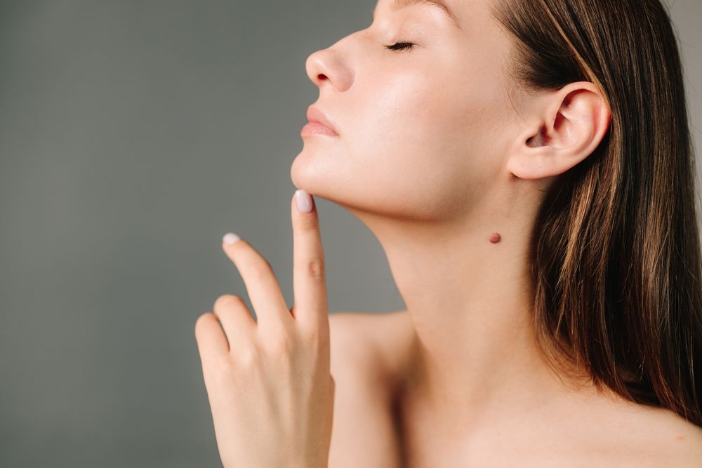 Woman with closed eyes, touches her chin with a finger, a mole visible on her neck, grey background.