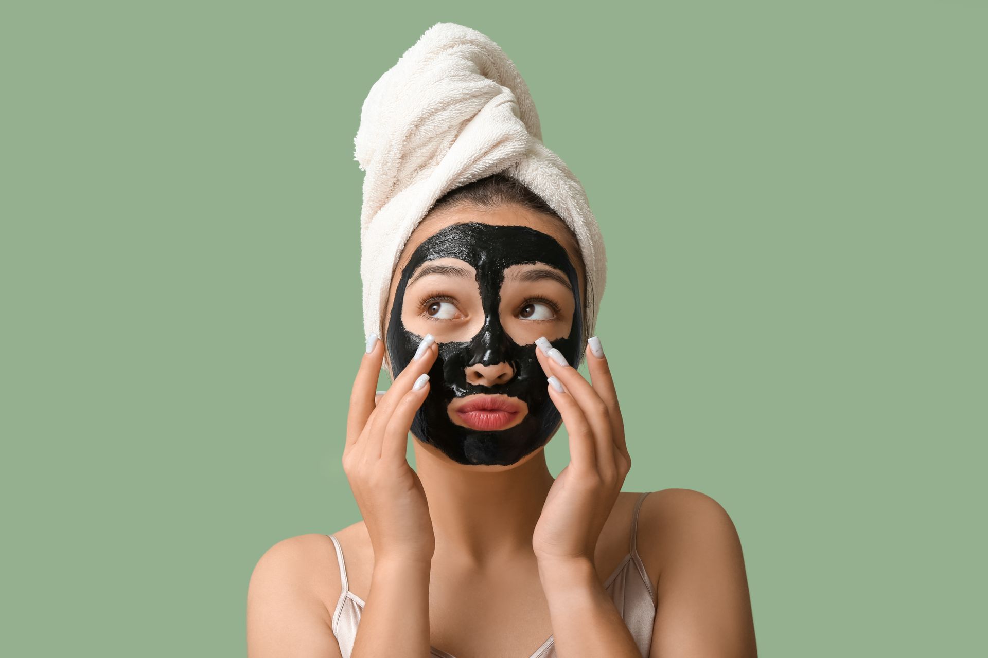 A woman with a towel wrapped around her head is applying a black mask to her face.