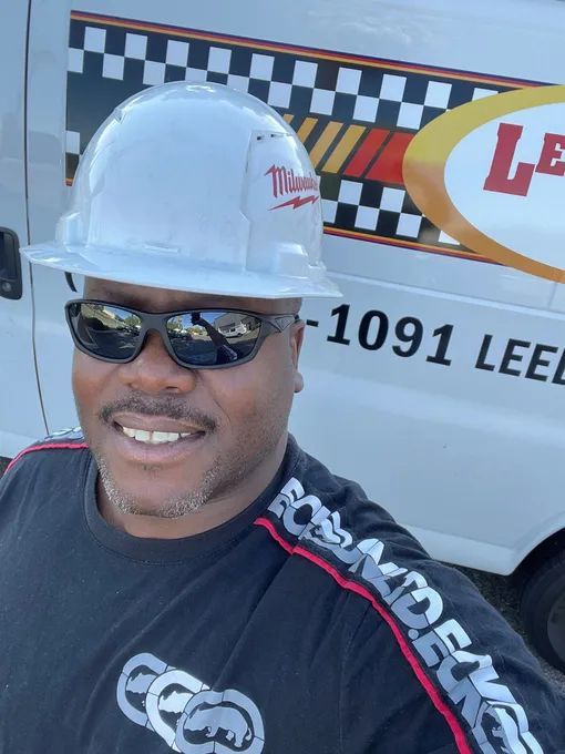 Man wearing a white hard hat and sunglasses smiles in front of a company van.