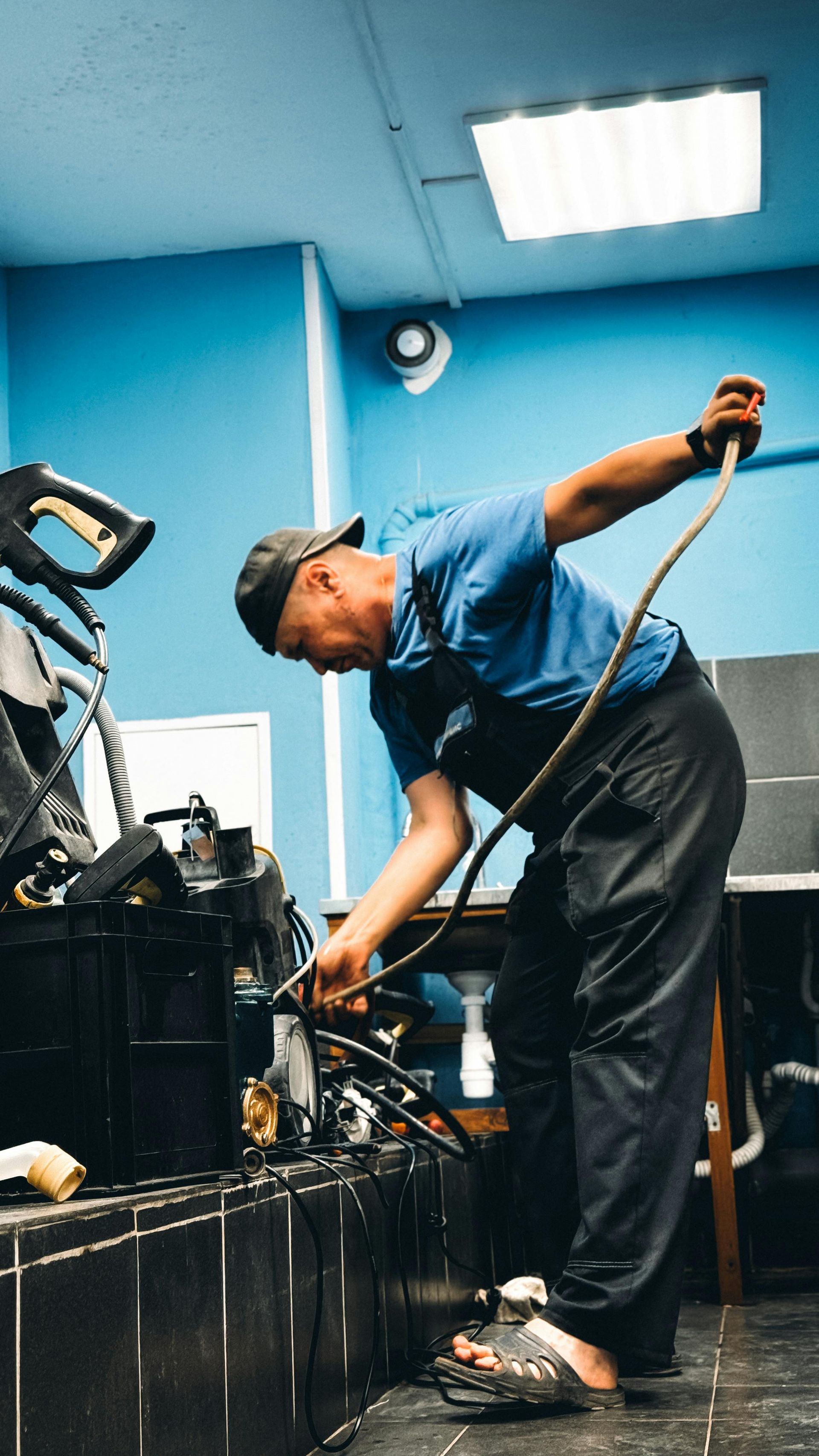 Mechanic in blue shirt working under a car in a garage, using a tool beside equipment and a lift