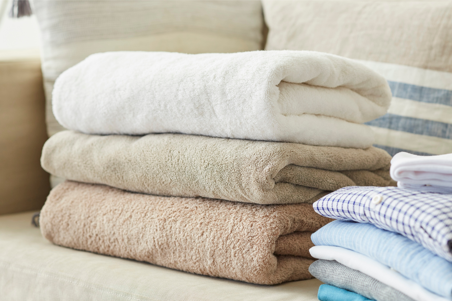 A stack of three folded towels—white, beige, and tan—resting on a sofa next to a stack of folded shirts.