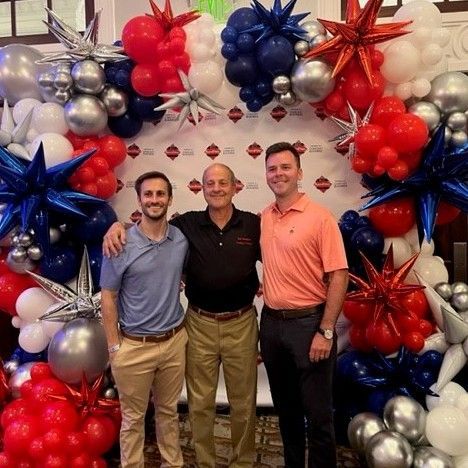 Three men are posing for a picture in front of a wall of balloons.