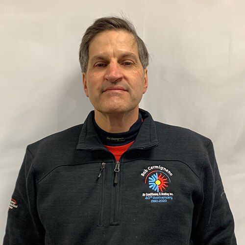 A man wearing a black jacket and a red shirt is standing in front of a white wall.