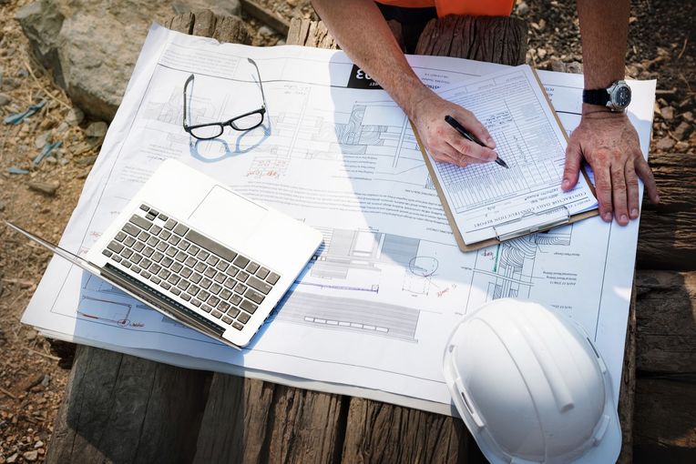 Architect reviewing blueprint plans outdoors with laptop, notebook, hard hat, and glasses on the table