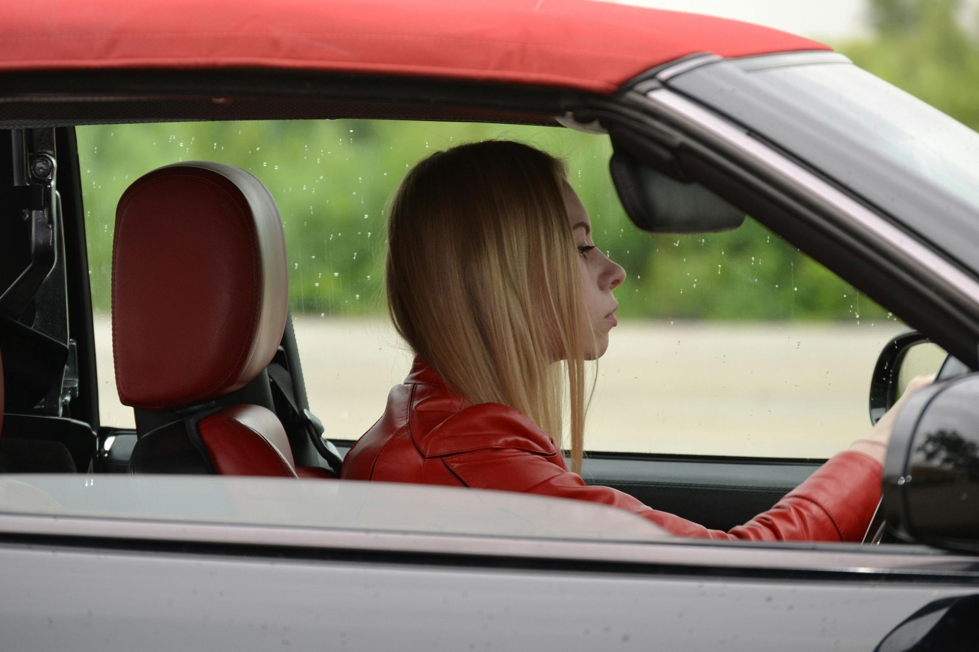 A woman in a red leather jacket is driving a black convertible car.