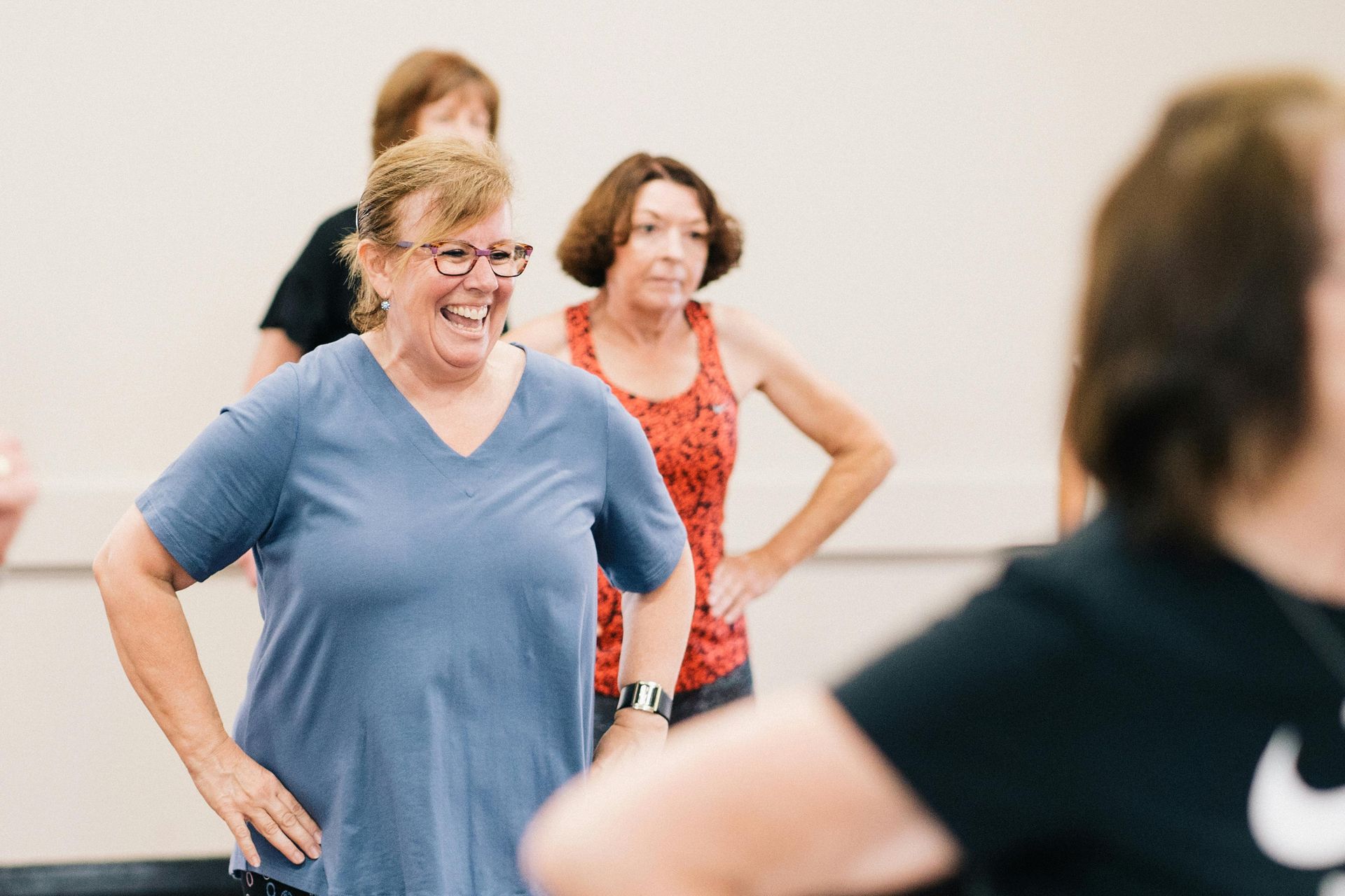 People participating in a fitness class in a bright indoor studio. A person in a blue shirt smiles while exercising.