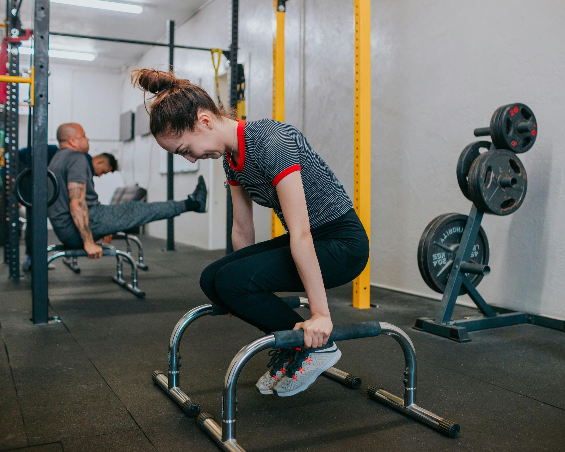 A person performs a tuck hold on parallettes in a gym, while another person exercises on a bench in the background.