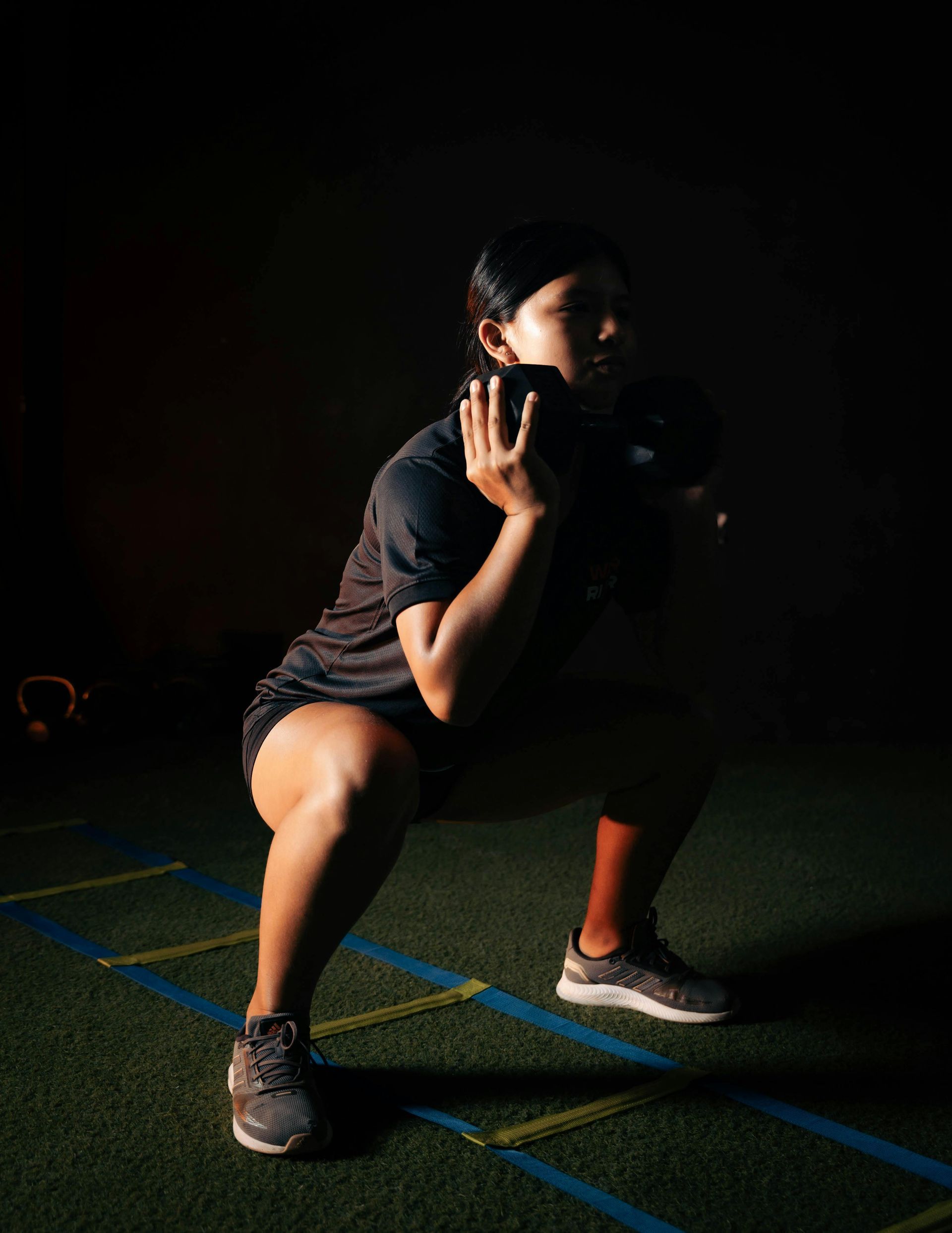 A person in dark gym attire performs a goblet squat while holding a dumbbell against their chest on an agility ladder.