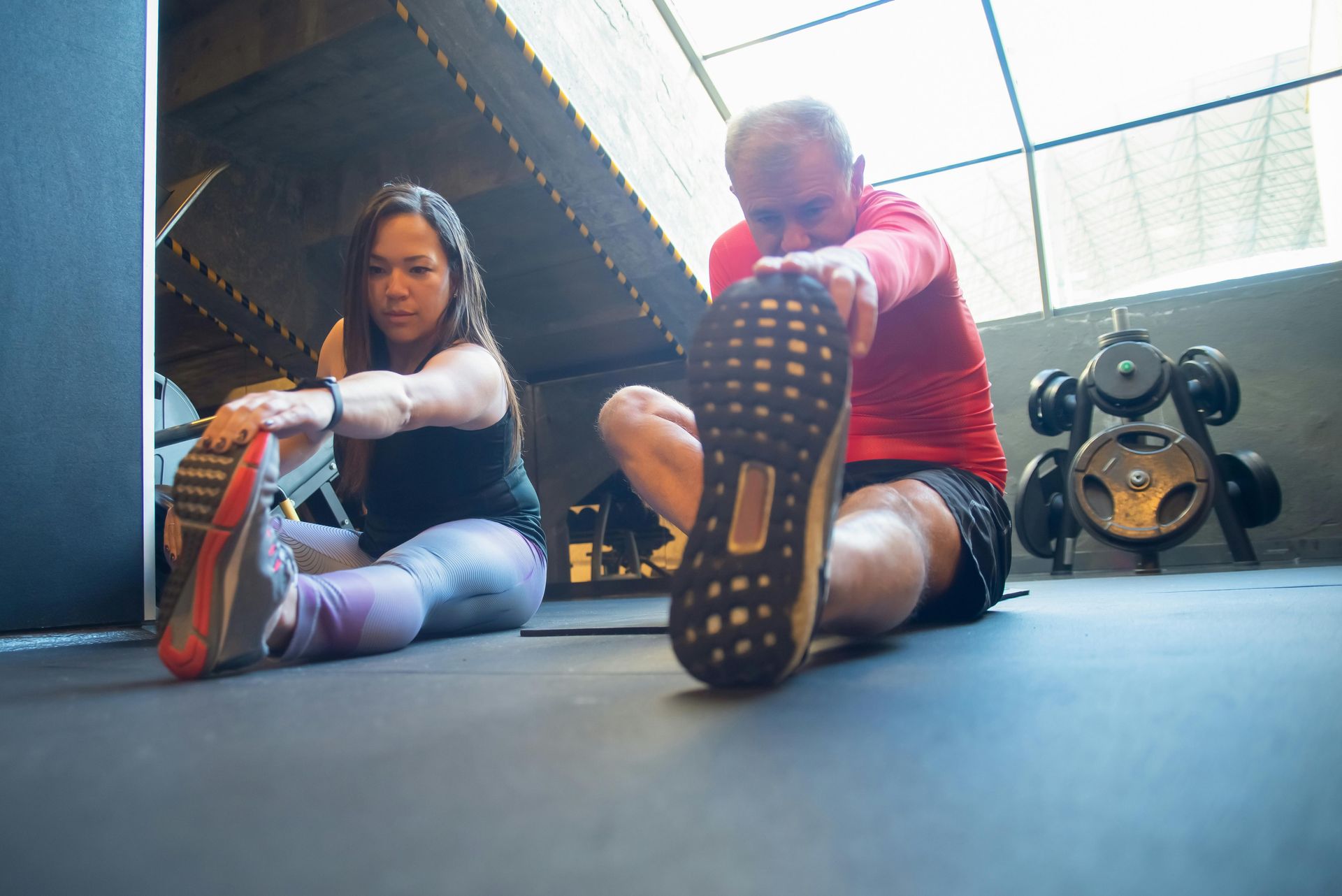 Two people sit on a gym floor, stretching their legs and reaching toward their toes in a sunlit workout space.