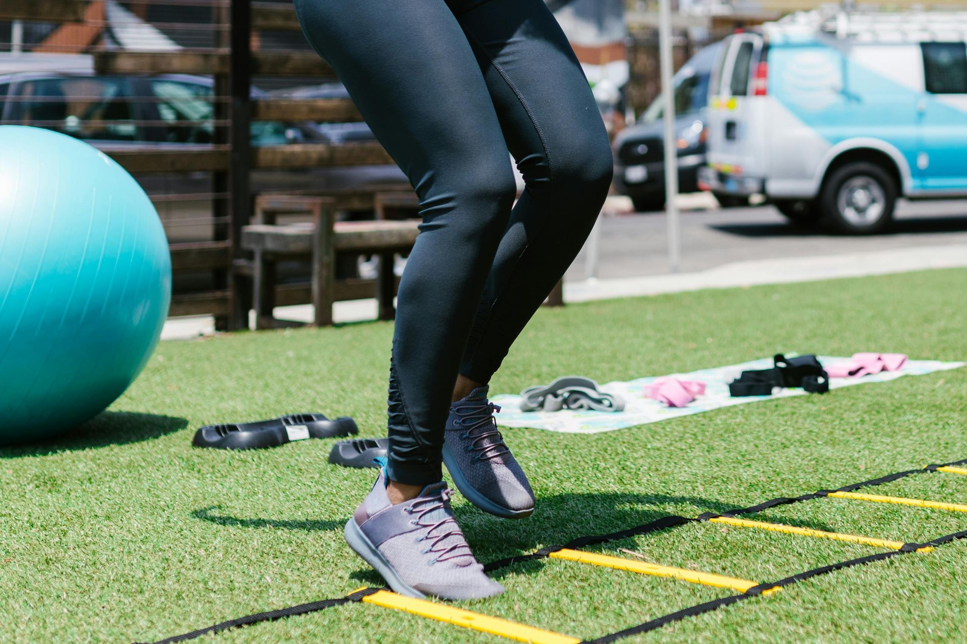 A person in black leggings performs agility ladder drills on artificial turf outdoors near gym equipment.