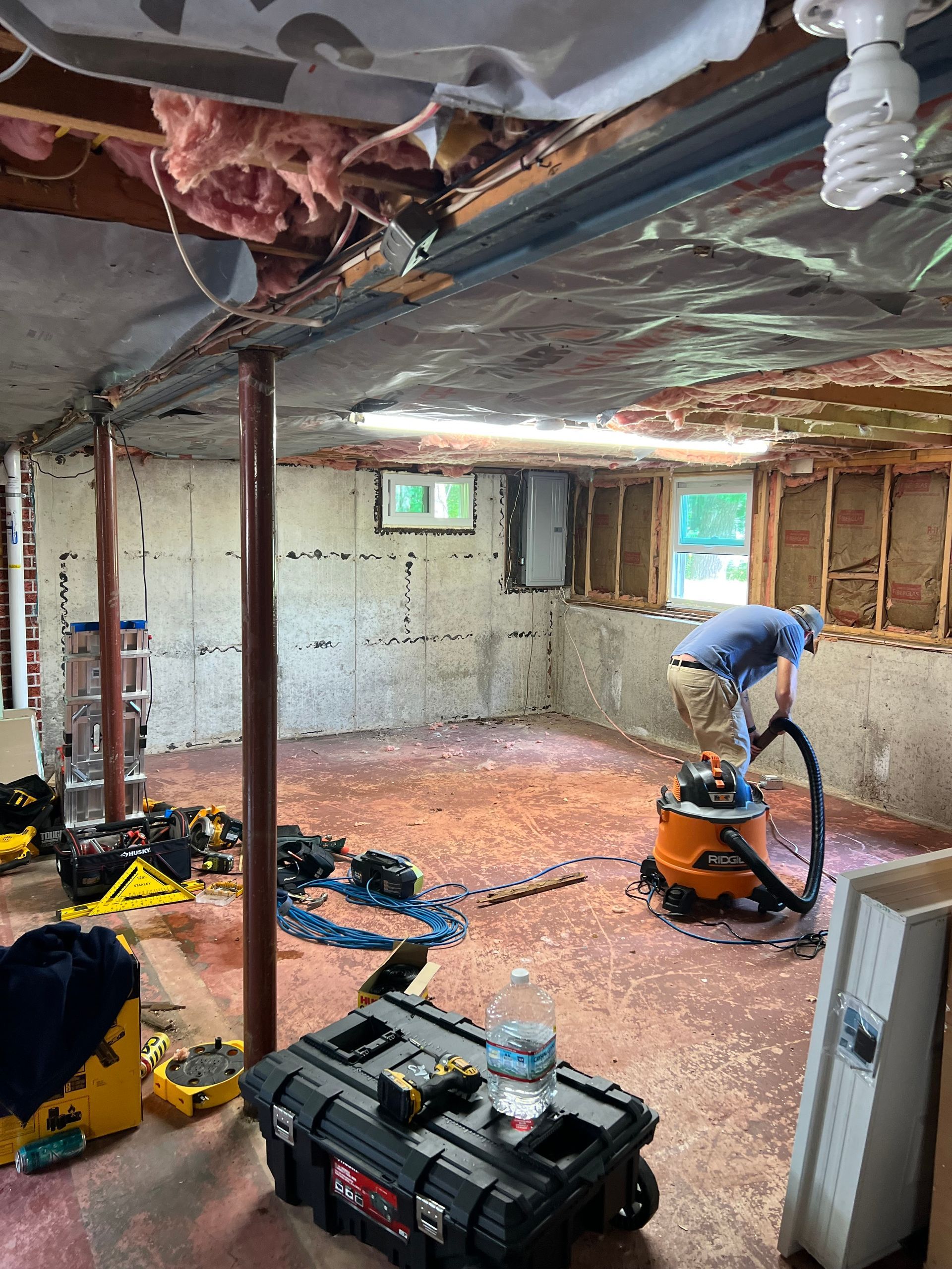 A person vacuums a dusty basement under construction. Exposed insulation and framing are visible.