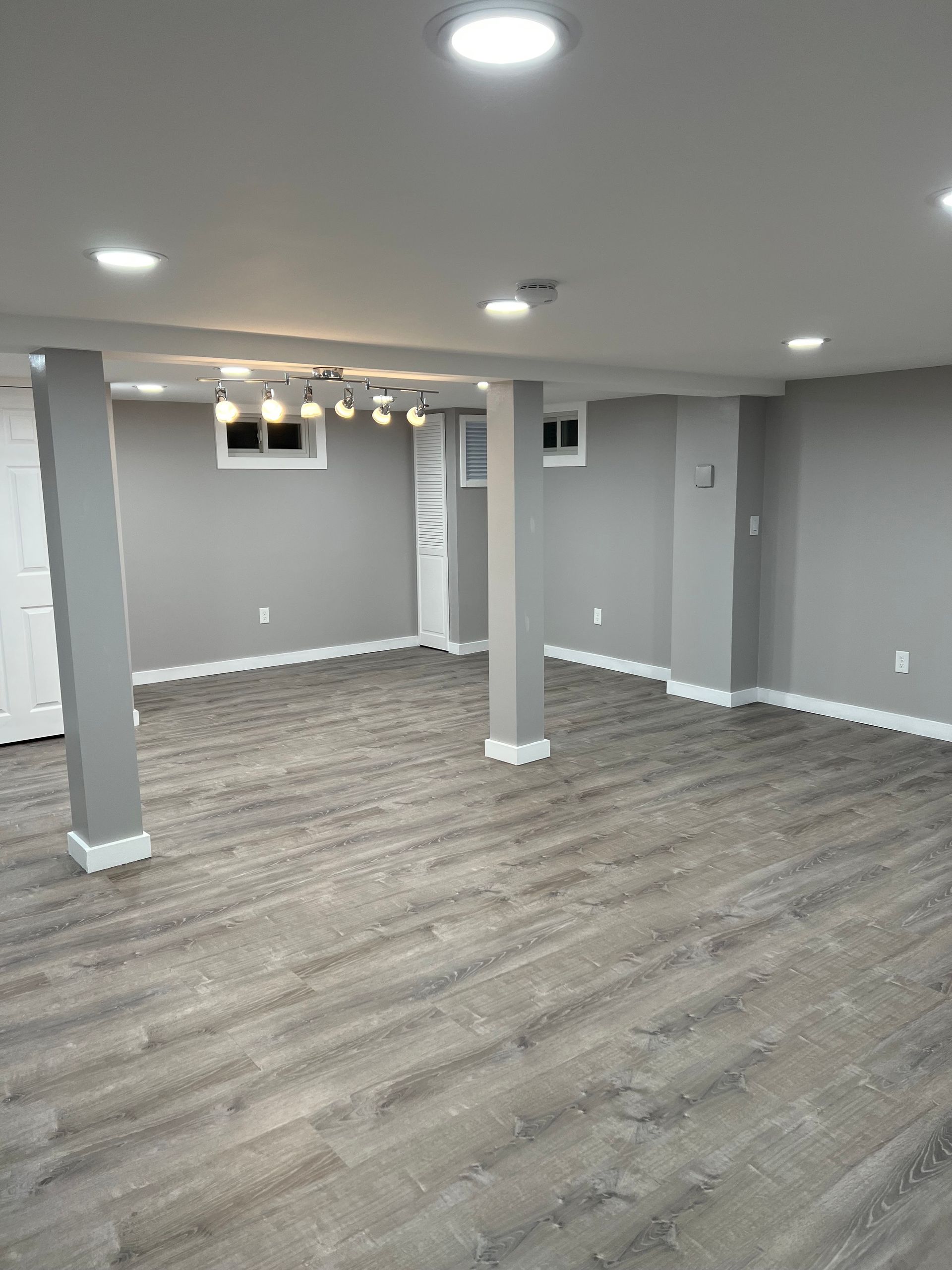 Empty basement with gray walls, flooring, and support columns. Overhead recessed lighting illuminates the space.