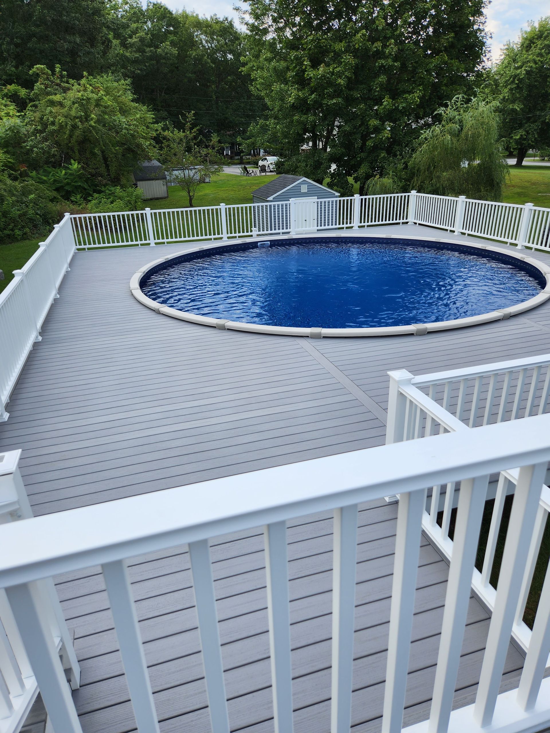 An above-ground, circular pool surrounded by a gray deck with white railings. The scene is outdoors with trees in the background.