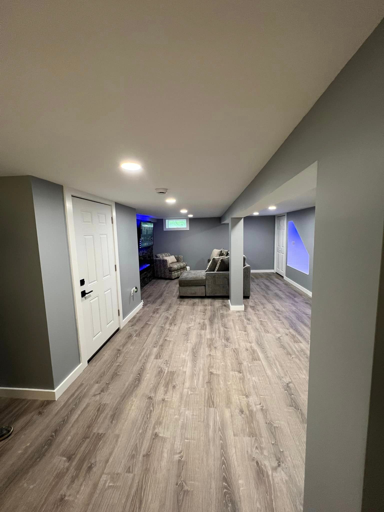 Finished basement with gray walls, wood-look flooring, recessed lighting, and a gray couch. A white door and doorway are visible.