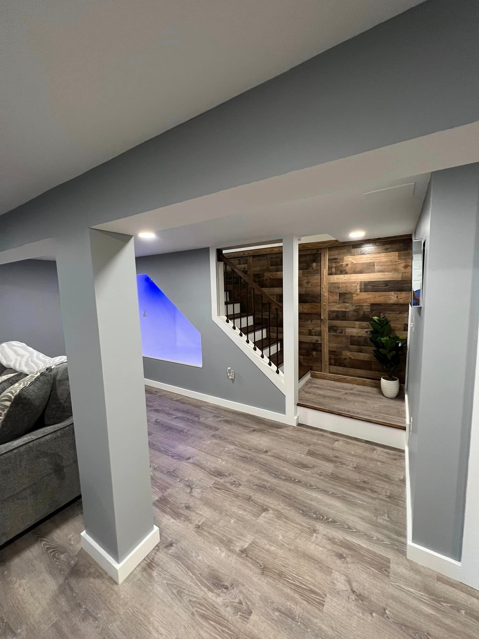 Finished basement with gray walls, wood-look flooring, and a staircase leading to a room with a wood-paneled wall.