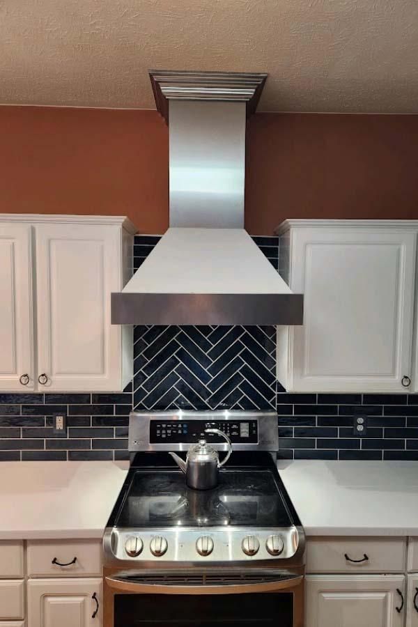 A stainless steel range hood above a stove, centered between white cabinets and a dark blue herringbone tile backsplash in a kitchen.