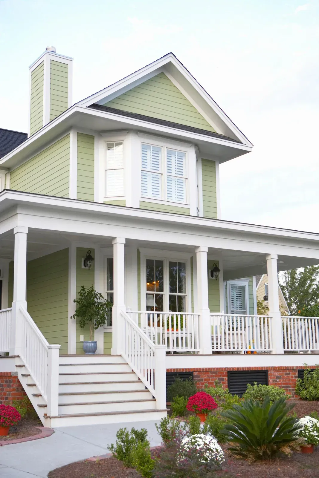 Two-story house with green siding, white porch, and brick foundation, surrounded by landscaping.