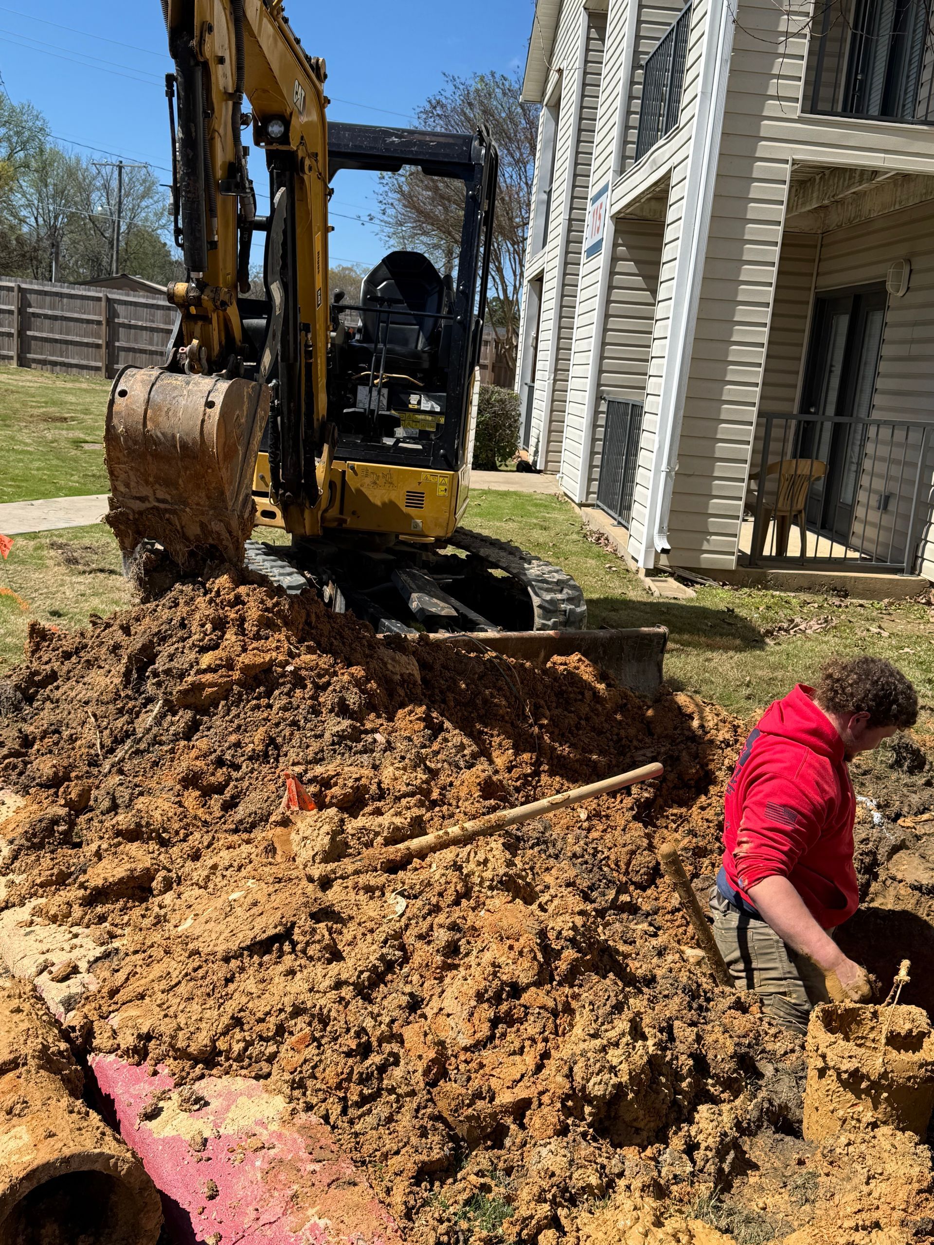 Backhoe digging a large trench beside a house while a worker in red kneels nearby