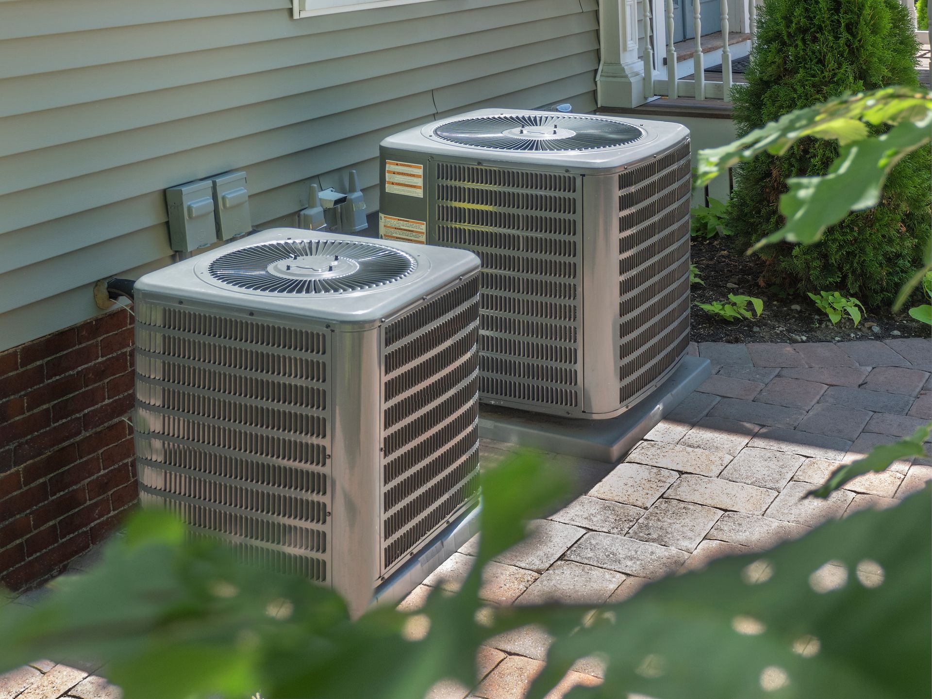 Two outdoor air conditioning units beside a house, partially framed by green leaves