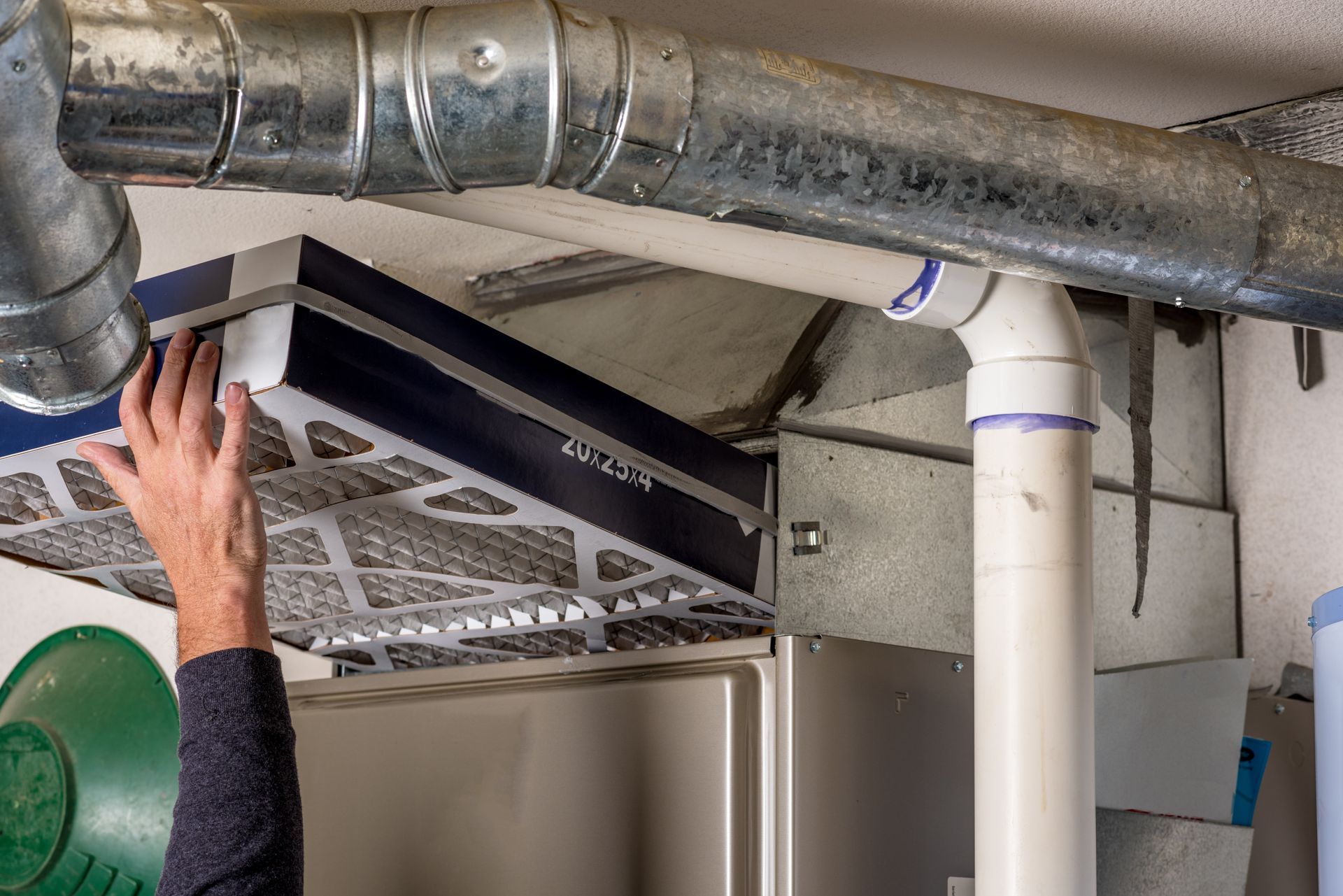 Hand adjusting a ceiling-mounted HVAC filter above a kitchen appliance near ductwork and pipes