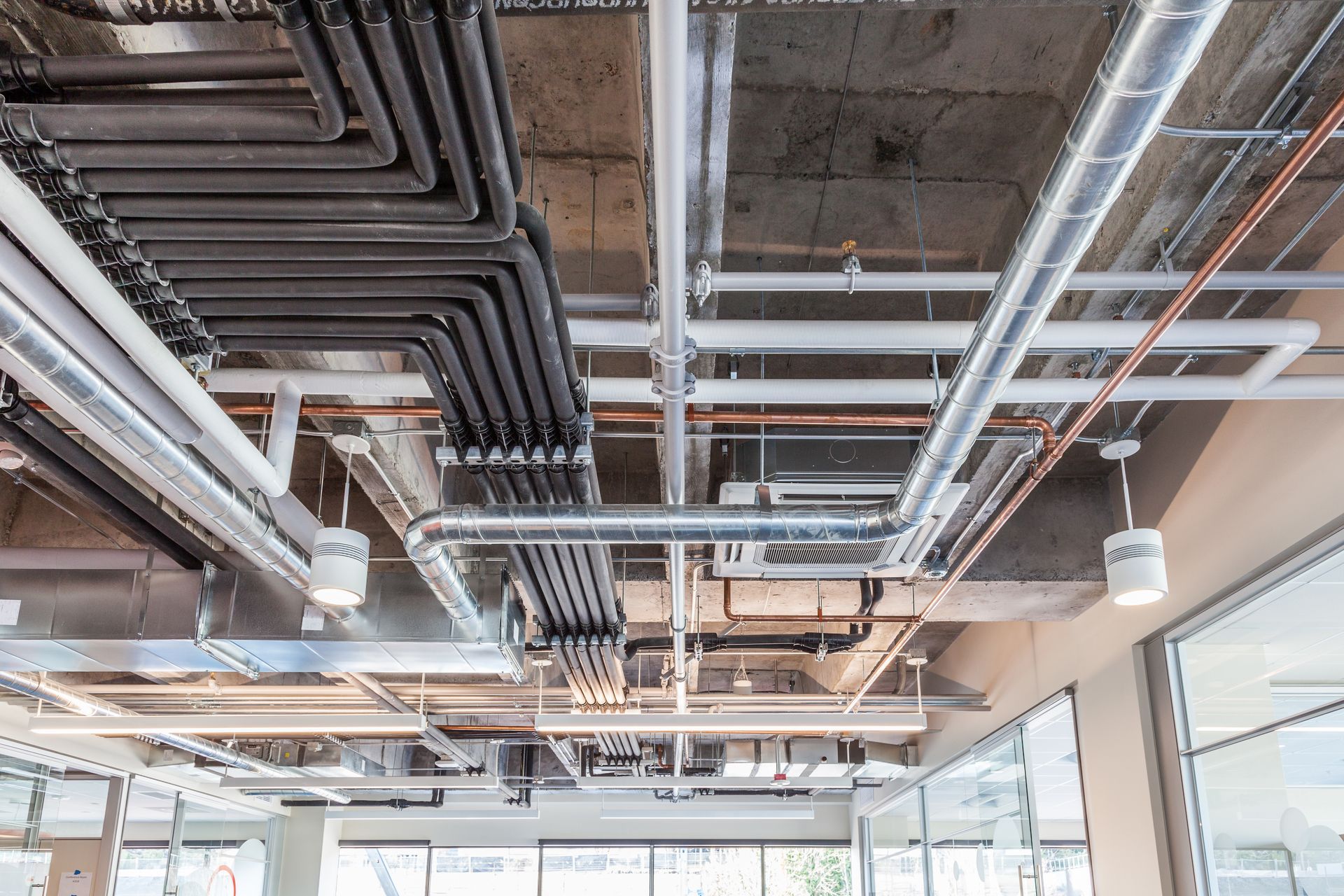 Exposed industrial ceiling with black pipes, silver ductwork, and hanging lights in a modern office space