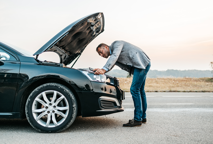 Man in jeans and a jacket leans over a black car with its hood open on a road, looking inside. | Walk's Towing