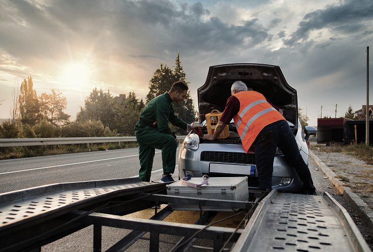 Two men loading a car onto a flatbed tow truck on a roadside. One wears green, the other an orange vest. | Walk's Towing