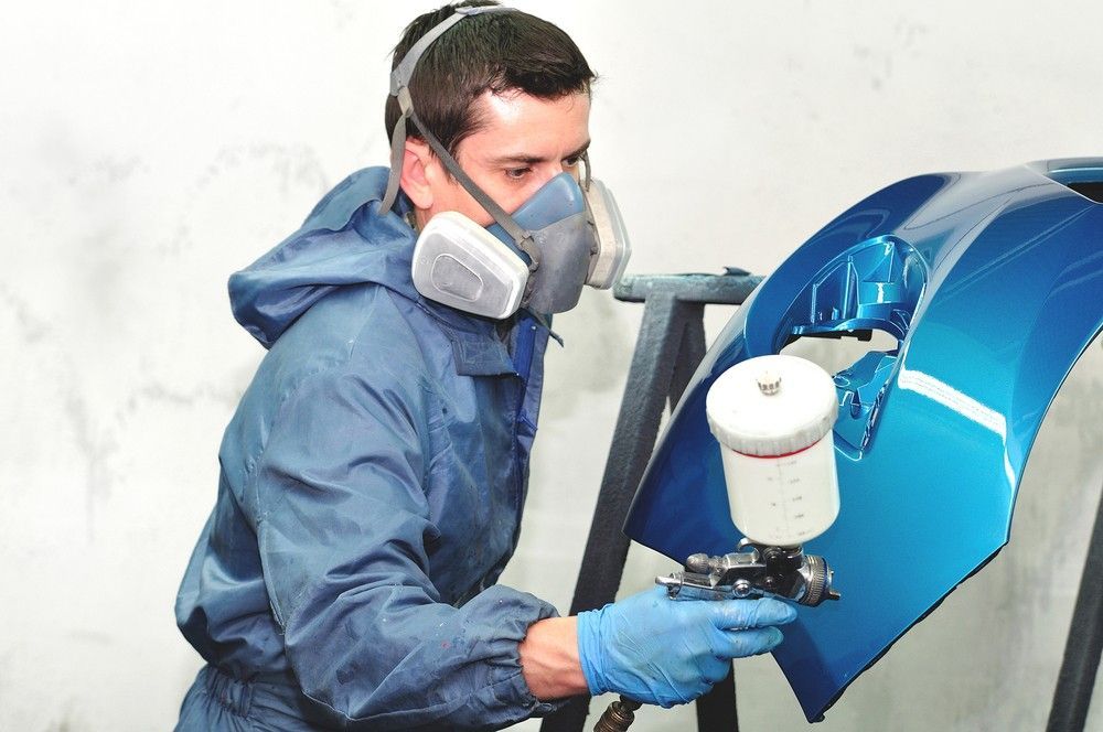 A Man Wearing a Mask and Gloves is Spray Painting a Blue Car Bumper — Dawson Crash Repair In Aitkenvale, QLD