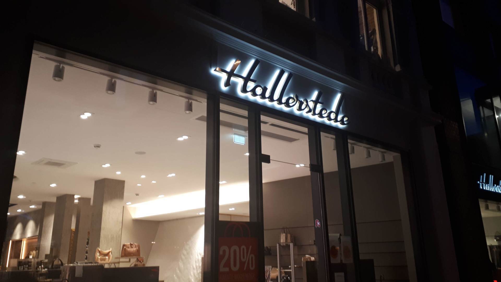 Hallhverste-Ladenfront bei Nacht; beleuchtetes Schild, große Glasfenster mit Innenauslage, 20 % Rabatt-Schild.