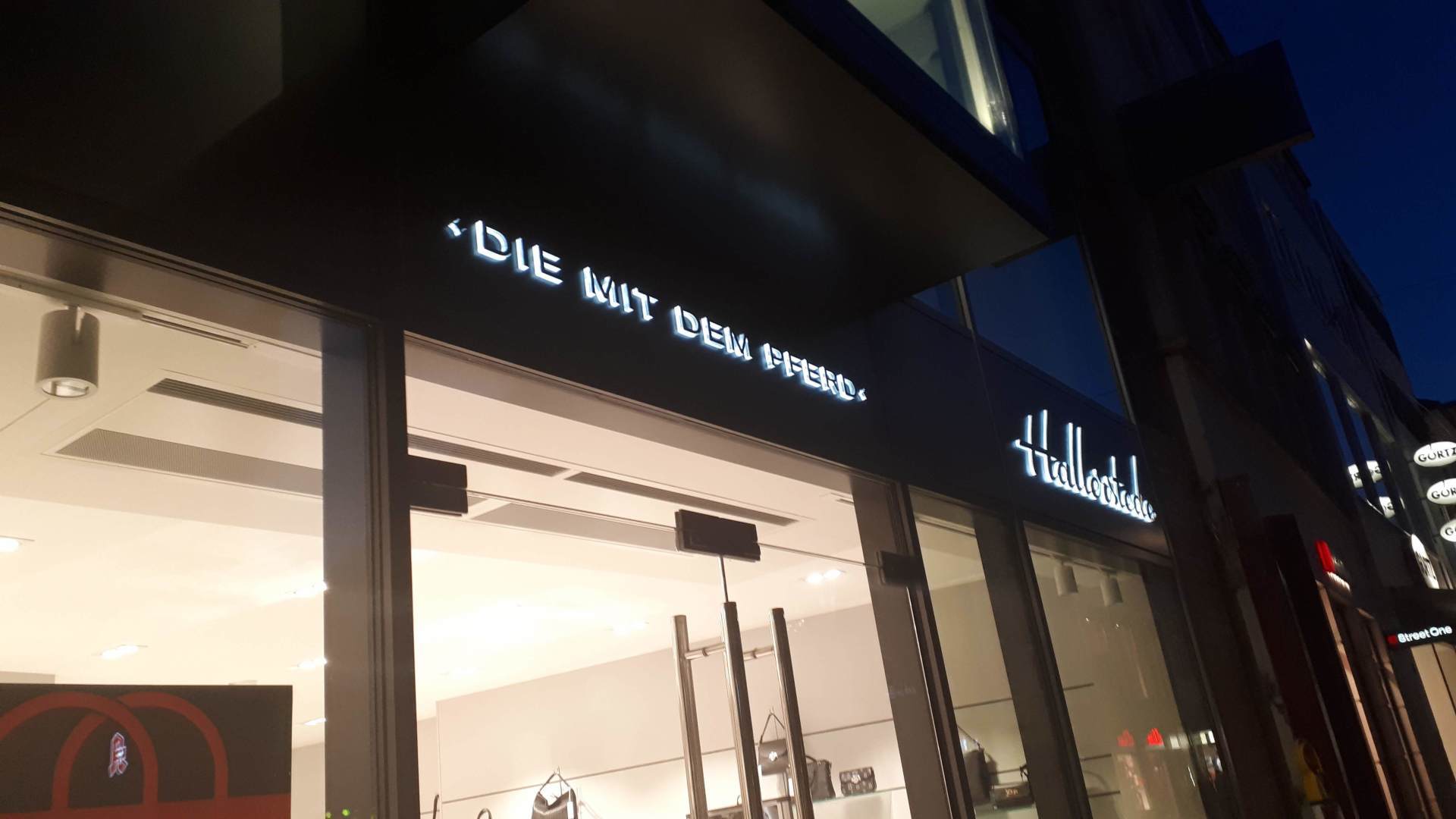 Ein Ladeneingang bei Nacht mit beleuchtetem Text auf Deutsch: „Die mit dem Pfeil“.