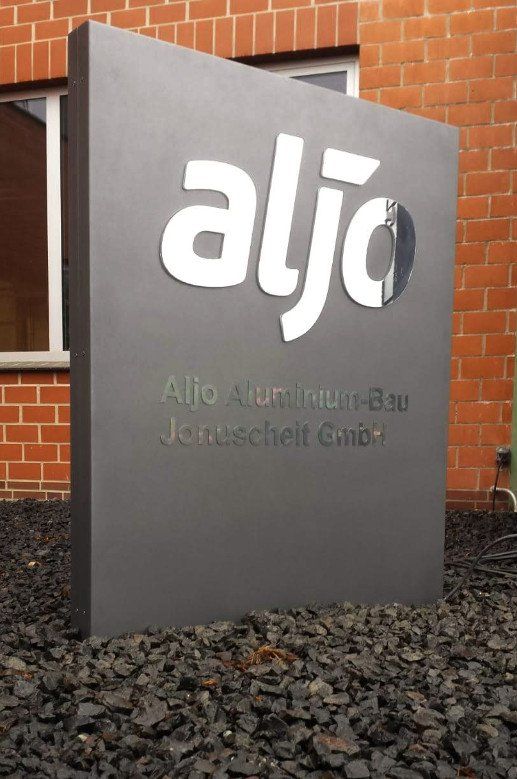 Graues Schild mit weißem „aljo“-Logo, Hintergrund Backsteingebäude, Schild für Aljo Aluminium-Bau.
